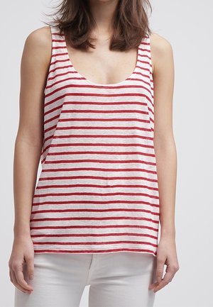 Kvinde iført ærmeløs hvid tanktop med vandrette røde striber og hvide bukser, stående foran en ensfarvet lys baggrund.