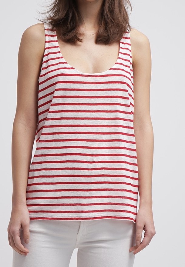 Žena v bezrukávovém bílém tričku s vodorovnými červenými pruhy a bílých kalhotách, stojí na pozadí v jednobarevné světlé barvě.