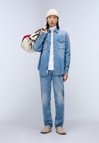 Lys blå denimskjorte med to frontlommer, parret med matchende jeans. Cremegul hue og beige sko, holder en flerfarvet taske.