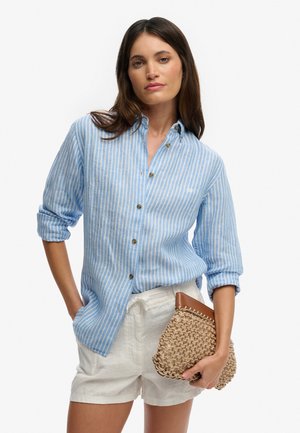 Kobieta ubrana w niebiesko-białą pasiastą koszulę zapinaną na guziki, białe szorty, trzyma plecioną beżową kopertówkę z brązową skórzaną obwódką.