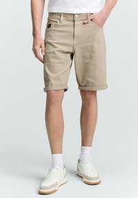 Beige farmernadrág sort, slim fit fazonnal, felhajtott szárakkal, elülső zsebekkel és egy kis címkével a csípőn. Fehér tornacipővel és zoknival párosítva.