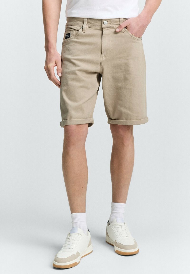 Beige farmernadrág sort, slim fit fazonnal, felhajtott szárakkal, elülső zsebekkel és egy kis címkével a csípőn. Fehér tornacipővel és zoknival párosítva.