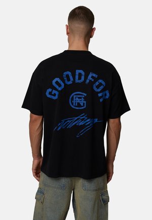 Mężczyzna o krótkich włosach, ubrany w czarną koszulkę z niebieskim napisem "GOODFOR" i grafiką, w połączeniu z wyblakłymi niebieskimi spodniami cargo, widziany od tyłu.