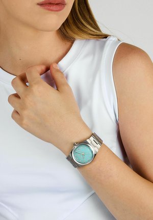 Orologio con cinturino argento e quadrante turchese, caratterizzato da indicatori delle ore puliti e una corona. Indossato su un polso chiaro con un indumento bianco.