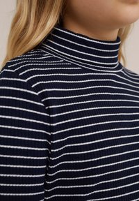 Pulover s vysokim ovratnikom s mornarskimi črtami in belimi vodoravnimi črtami. Ribana tekstura, tesno prileganje in podrobnost visoke ovratnice so opazne.