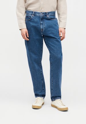 Homme portant un jean bleu à jambes droites, une chemise beige à manches longues et des baskets blanches, debout devant un fond uni.