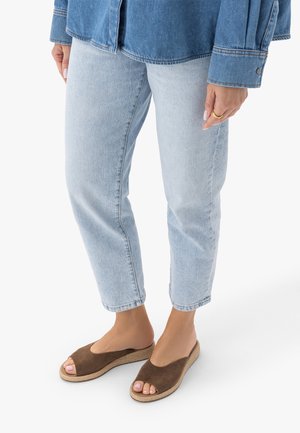 Osoba ubrana w jasnoniebieskie dżinsy, kurtkę dżinsową oraz brązowe, otwarte, wsuwane sandały, stojąca na białym tle.