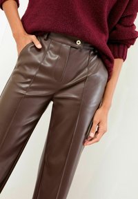 Smeđe hlače od umjetne kože s ravnim prednjim dijelom, vertikalnim šavovima i srebrnim kopčanjem. Uparene s udobnom bordo pulovericom.