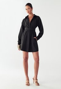 Must mustrõivad, millel on pikad varrukad, vöökoht, krae ja küljetaskud. Sile kanga; kombineeritud beežide kontsakingadega.