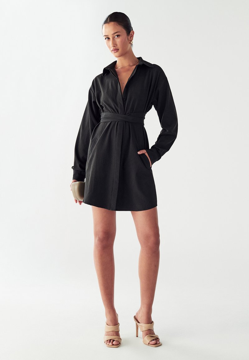 Must mustrõivad, millel on pikad varrukad, vöökoht, krae ja küljetaskud. Sile kanga; kombineeritud beežide kontsakingadega.