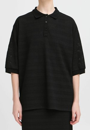 Personne portant un polo noir ample à manches courtes avec des motifs horizontaux subtilement texturés et trois boutons sur la patte avant.