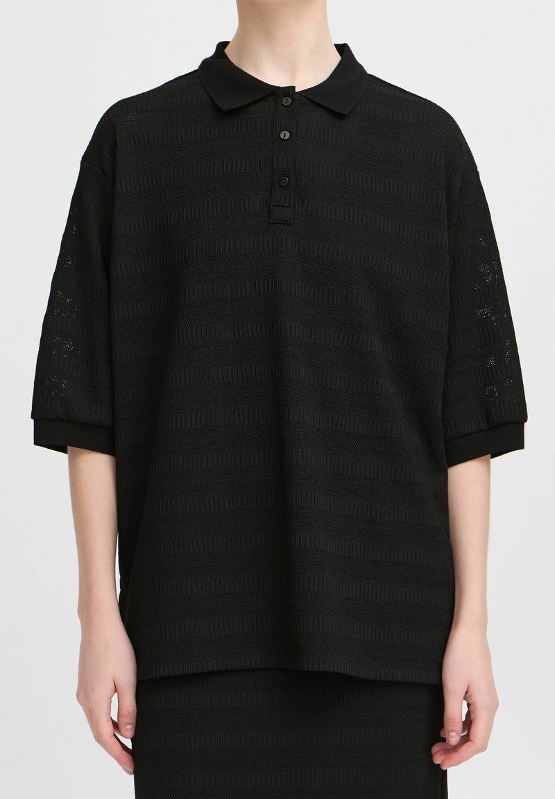 Personne portant un polo noir ample à manches courtes avec des motifs horizontaux subtilement texturés et trois boutons sur la patte avant.