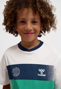 Göndör hajú gyerek mosolyog, fehér, tengerészkék és zöld Hummel pólót visel "Változtasd meg a világot a sporton keresztül" felirattal.