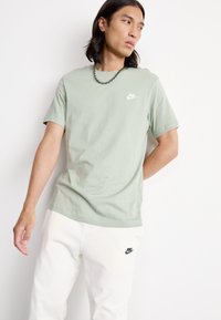 T-shirt de algodão verde-claro com decote redondo e mangas curtas. Apresenta um pequeno logotipo branco da Nike no lado esquerdo do peito. Usado com calças de treino brancas.
