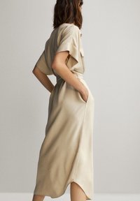 Beige midi-mittainen mekko pehmeästä kankaasta, jossa lyhyet hihat, vyötäröllä vyö, sivutaskut ja laskeutuva siluetti, jossa on pieni sivuhalkio.