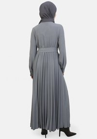 Langes, gefaltetes graues Kleid mit langen Ärmeln und einem taillierten Bund. Es hat ein fließendes Design und eine glatte Textur, kombiniert mit schwarzen High Heels.