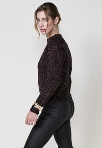 Dunkelbrauner gestrickter Pullover mit mehrfarbigen Fäden, rundem Halsausschnitt und geripptem Saum. Getragen mit schwarzen, eng anliegenden Hosen und einem Metallarmband.