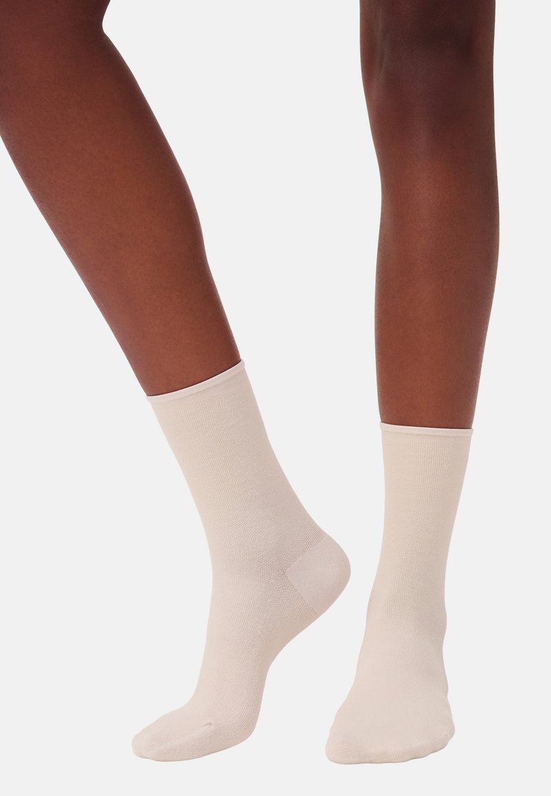 Beige Baumwollsocken mit einem gerippten Bündchen, glatter Textur und verstärkter Ferse. Die Socken werden an Füßen mit dunklem Hautton getragen.