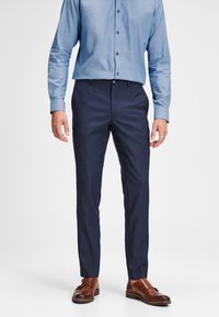 Pantalons navy à jambe droite dans une coupe ajustée, associés à une chemise à manches longues bleu clair. Des chaussures en cuir marron complètent l'ensemble.