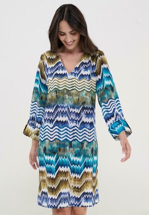 Mujer sonriente mirando hacia abajo, vestida con un vestido de manga larga y escote en V con patrones de zigzag en azul, verde, blanco y marrón.