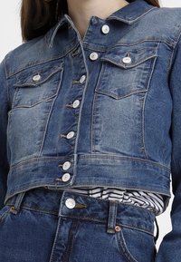 En kortad denimjacka i medium blå med en urblekt finish. Har två bröstfickor och silverfärgade knappar. Strukturerad tyg; knappstängning framtill.