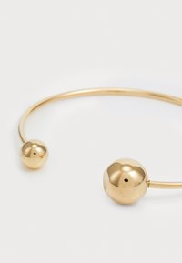 Guldmetall cuffarmband med en slät, rundad design som har två sfäriska accenter i vardera änden. Snygg, polerad yta.