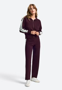 Bordo športni komplet z jopico z zadrgo in ujemajočimi hlačami. Svetleče stranske črte, prilegajoč kroj in mehka tekstura tkanine.