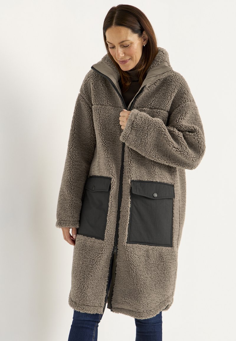 Flauschiger beiger Mantel mit Reißverschluss vorne, großen grauen Taschen und Kapuze. Verfügt über einen weichen, strukturierten Stoff und eine lockere Passform.
