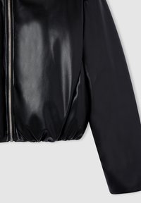 Czarna, błyszcząca kurtka z sztucznej skóry z przednim srebrnym zamkiem błyskawicznym i ściąganym dołem na jasnoszarym tle.