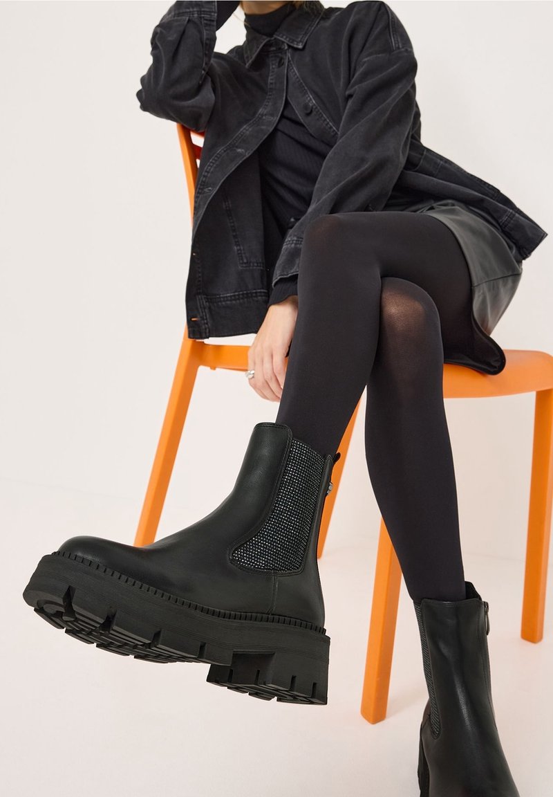 Bottines en cuir noir avec une semelle épaisse, panneau latéral élastique et détails texturés. Portées avec des collants noirs, assise sur une chaise orange.
