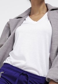 Weißes V-Ausschnitt-T-Shirt unter einem grauen Blazer, kombiniert mit lila Jogginghosen mit Kordelzug. Der Stoff wirkt weich und hat eine lockere Passform.