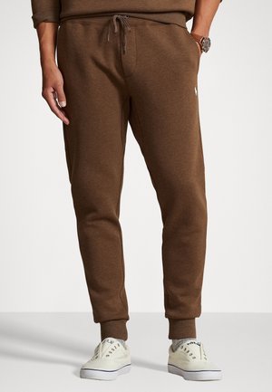 Mann trägt braune Jogginghosen, ein passendes braunes Sweatshirt, graue Socken, weiße Sneakers und eine Armbanduhr vor einem einfarbigen Hintergrund.