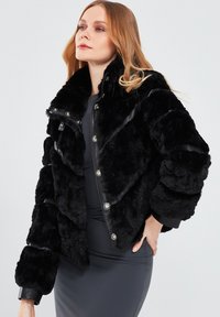 Black faux fur jacket with textured surface, quilted design, snap buttons, and ribbed cuffs. Features a high collar and a cozy fit.