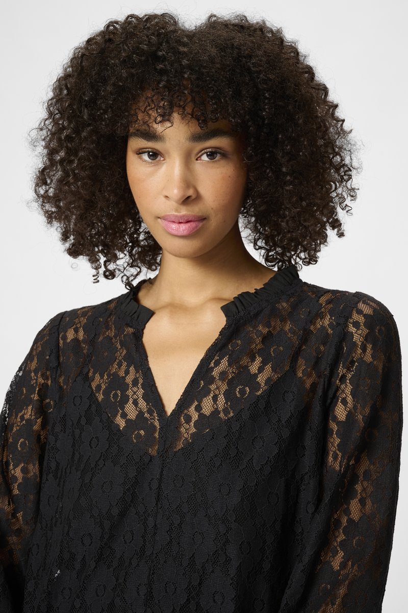 Blusa de encaje negro con patrones florales, escote en V pronunciado y cuello con volantes. La tela es semitransparente y tiene mangas largas.