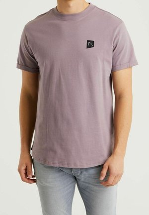 Mężczyzna noszący stonowany fioletowy t-shirt z krótkim rękawem, z małym czarnym kwadratowym logo na lewej piersi, w połączeniu z jasnoszarymi dżinsami.