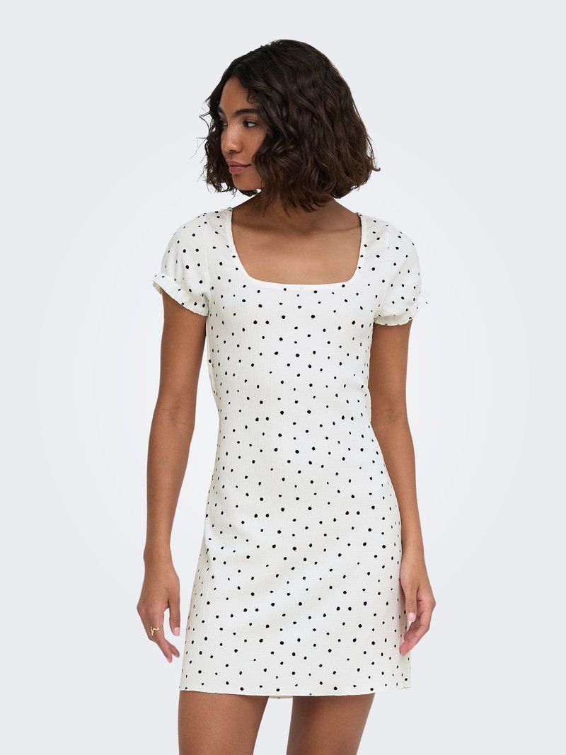 Femme aux cheveux bouclés mi-longs porte une robe mini blanche à manches courtes avec des pois noirs, regardant vers sa gauche sur un fond uni.