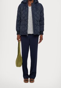 Marineblå quiltet dunjakke med hætte, grå strikket sweater, marineblå bukser og en grøn ruskind skuldertaske, kombineret med brune sko.