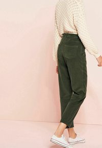 Grüne Cordhosen mit hoher Taille, schmalem Bein und zwei Gesäßtaschen, kombiniert mit einem strukturierten cremefarbenen Pullover und weißen Sneakers.
