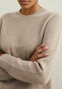 Pulover din lână bej cu guler rotund, manșete drepte și textură subtilă. Prezintă o croială lejeră, cu detalii vizibile ale cusăturilor pe mâneci.