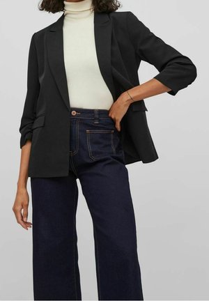 Femme portant un blazer noir sur un turtleneck blanc rentré dans un jean bleu foncé taille haute à jambes larges, une main sur la hanche.