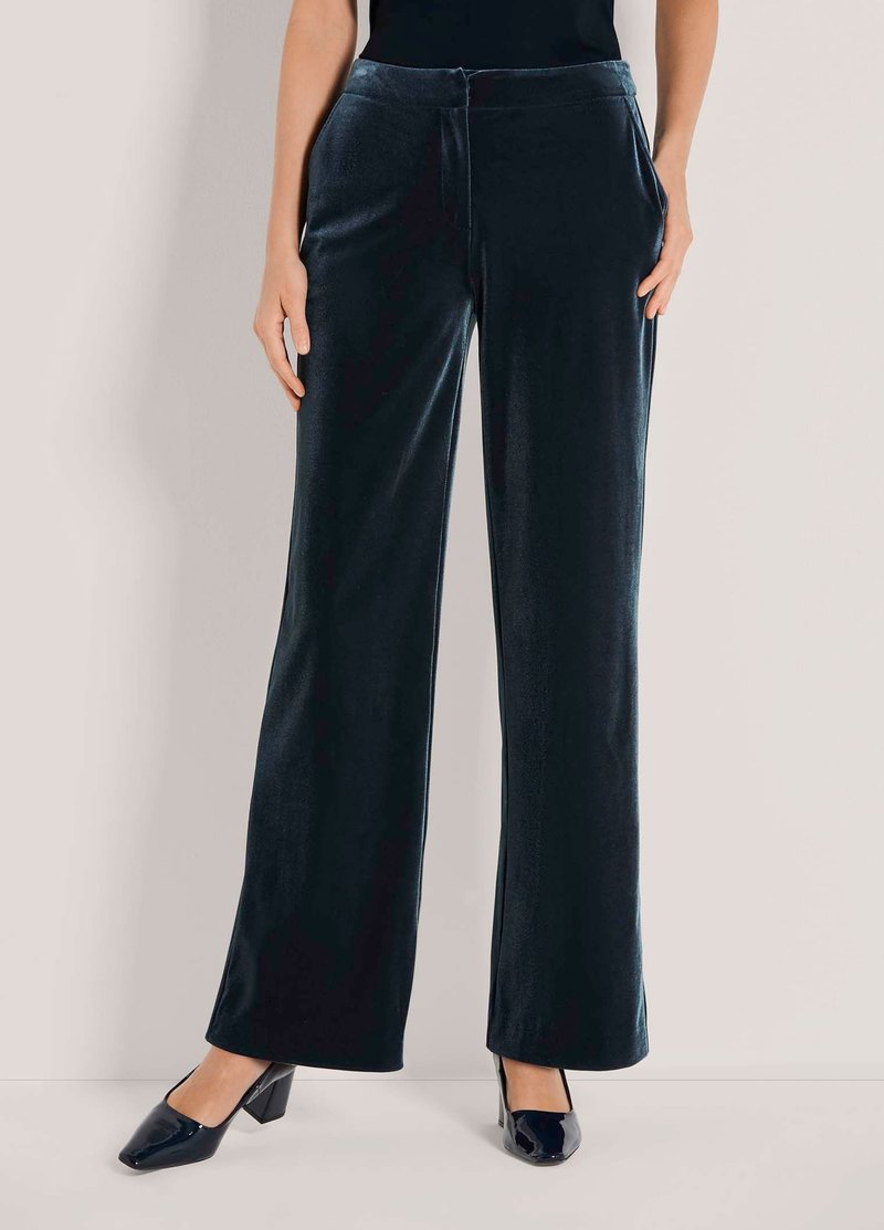 Vrouw in hooggemeten, wijdvallende zwarte fluwelen broek, gecombineerd met zwarte blokhakken, staande tegen een effen achtergrond.