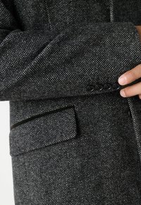 Szürke textúrájú anyagú blézer, amely slim fit fazonnal rendelkezik, és három látható fekete gomb található az ujjon, valamint egy diszkrét zseb az oldalon.