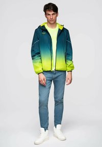 Mladý muž stojící v modro-zelené gradientové kapucí, neonově žlutém tričku, modrých džínách a bílých vysokých teniskách na bílém pozadí.