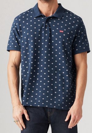 Mand iført en marineblå Levi's poloshirt med små hvide krone-mønstre og mørkeblå jeans, stående med afslappede arme.