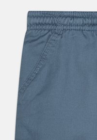 Hellblaue Hose mit elastischem Bund, diagonal angeordneten Fronttaschen und einem weichen, strukturierten Stoff. Sichtbare Nähte.