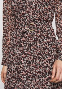 Robe fleurie avec de petites fleurs roses, blanches et noires, dotée d'une ceinture en tissu avec une boucle ronde dorée et des boutons noirs sur le devant.