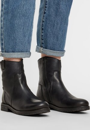Bottines en cuir noir avec une finition lisse, fermeture éclair sur le côté et talon bas, associées à un jean en denim bleu retroussé.