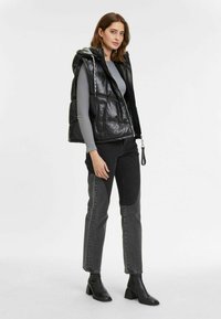 Schwarze gepolsterte Weste mit Kapuze, glänzender Oberfläche, Fronttaschen und Reißverschluss, getragen über einem grauen Langarmshirt und einer schwarzen Jeans.
