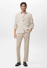 Chemise beige à boutons avec une poche poitrine, associée à un pantalon beige. La tenue présente une coupe décontractée et une texture lisse, complétée par des chaussures noires.