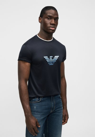 Άνδρας που φοράει μπλε κοντομάνικο πουκάμισο με λογότυπο αετού σε ανοιχτό μπλε διαβάθμιση και casual μπλε τζιν, στέκεται μπροστά σε απλό φωτεινό φόντο.